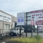Outdoor commercial area with large Japanese billboards on a building; a white van is parked near a chain-link fence in the foreground.